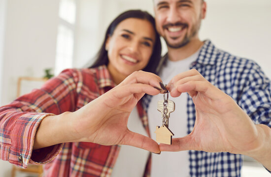 Happy young couple holding a key in hands in the form of heart standing at home looking at camera enjoying real estate purchase, smiling and celebrating moving day. Relocating, move, rent concept.