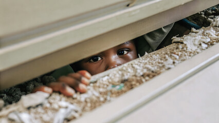 A young African boy peers out cautiously from under a crack.A young boy's gaze, drawn to something beyond the opening.Hidden observation, a child's face framed by a gap in the structure.
