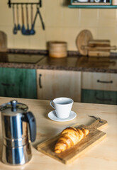 Warm light and cozy atmosphere in the kitchen. Coffee pot and hot coffee, empty cup and croissant on a wooden board.