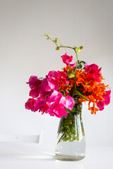 A vibrant bouquet of orange campis flowers and raspberry-hued bougainvillea rests on a white table, its reflection shimmering in a mirror. Ample space for text.