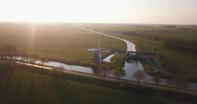 Summer morning sun opposite. Drone moves towards windmill. In the center of the frame is an old Dutch mill located on a green field near a canal with water. Province of Groningen. North Netherlands.