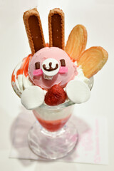 A cute dessert in a glass, resembling a bunny with pink ice cream, biscuits as ears, marshmallows, and strawberries, served in Japan, in a maid cafe.