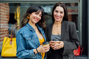 Two businesswomen standing in front of a shop window, holding takeaway coffee and smiling