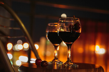 Two elegant glasses filled with dark liquid sit on a wooden table, overlooking a city illuminated at night. Cherries add a touch of color, enhancing the romantic atmosphere.