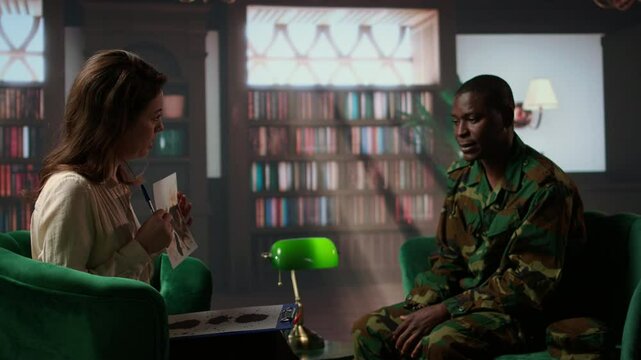 Female therapist presents a Rorschach inkblot test to a black soldier in armed forces uniform during a counseling session. Diverse people at therapy in a warm office with bookshelves. Camera A.