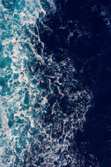 Aerial shot capturing the contrast between dark ocean waves and bright white seafoam, creating dynamic and dramatic seascape. Backgrounds or marine themes