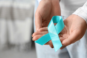 Young woman holding turquoise ribbon at home, closeup. Cervical Health Awareness Month