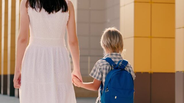 Loving mother accompanies little son on way to educational institution. Young mother takes son with blue backpack to school holding hand. Caring mother helps son to get ready for school day