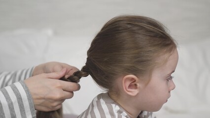 Mother braids professionally daughter brunette hair into hairstyle. Careful parent in children room...