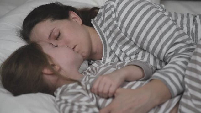 Brunette mother and little child sleep bed together. Mom and daughter lean cheeks hugging tightly. Woman and kid see sweet dreams cozy bedroom. Happy family. Children sleep with plush toy and mommy.