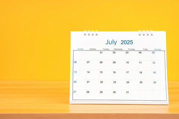 July 2025, Monthly desk calendar for 2025 year on wooden table on yellow background.