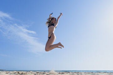 Obraz premium Mujer joven saltando de emoción porque esta feliz y divertida, con un sensacional día de vacaciones en verano en la playa, soleado con cielo azul.