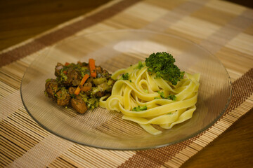 A delicious dish of tagliatelle pasta garnished with broccoli, served alongside sautéed chicken and mixed vegetables, presented on a glass plate with a wooden placemat backdrop.