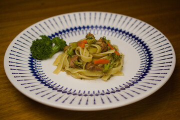A delightful dish of tagliatelle pasta, sautéed chicken, and fresh vegetables. Served with a broccoli garnish on a decorative plate featuring a blue and white design.