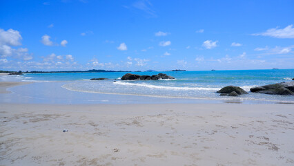 Fototapeta premium A pristine tropical beach with clear turquoise water, soft white sand, and large granite rocks. The sky is clear blue with scattered white clouds.