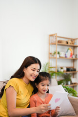 young asian mother accepting the little daughter hand painted card, little girl holding card for mom, sitting on the sofa in the living room, happy looking at each other, copy space for mother's day.