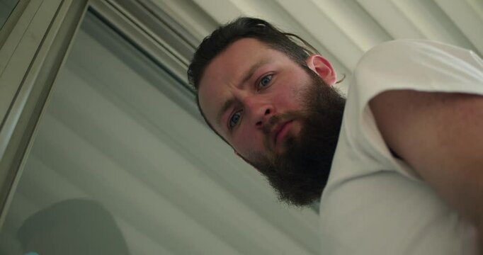 A low-angle shot captures a man with a beard and a white shirt, his face tense and curious as he looks downward. The perspective evokes a sense of unease or confrontation.