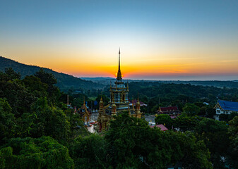 Fototapeta premium A breathtaking aerial view of a golden Buddhist temple at Wat Mae Klang Waterfall in Chiang Mai Thailand is surrounded by lush greenery, with the sunrise casting warm hues across the horizon.