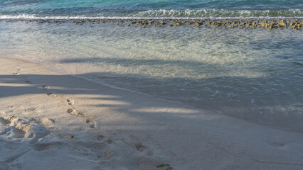 A chain of footprints of a man's bare feet runs on the sandy beach along the edge of clear turquoise water. Volcanic rocks are visible in the ocean. Mauritius.