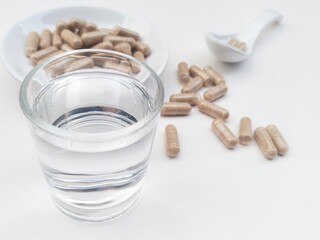 Capsule pills in a small white plate and in a white spoon, as well as a glass of water on a white background.