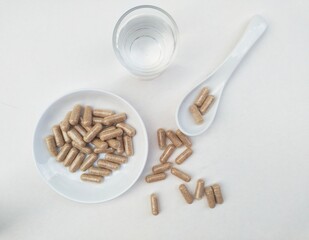 Capsule pills in a small white plate and in a white spoon, as well as a glass of water on a white background.