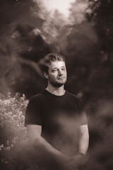 A sepia-toned portrait of a contemplative man in a natural setting. The soft-focus foreground and dappled light create a dreamy, reflective atmosphere, enhancing the artistic mood of the image