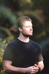 A handsome man in a black shirt stands surrounded by lush greenery, bathed in soft natural light. The background blurs with vibrant foliage, creating a peaceful and serene atmosphere