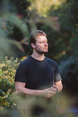 A handsome man in a black shirt stands surrounded by lush greenery, bathed in soft natural light. The background blurs with vibrant foliage, creating a peaceful and serene atmosphere