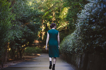 A woman walks confidently down a path in a lush green forest, wearing a dark green dress and black boots. Natural sunlight filters through the trees, creating a serene and vibrant atmosphere