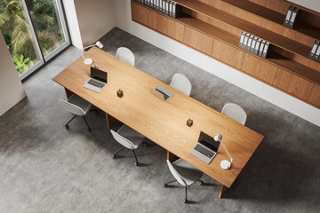 Modern office interior with wooden table, chairs, and laptops in open space with large windows and bookshelves in background. 3D Rendering