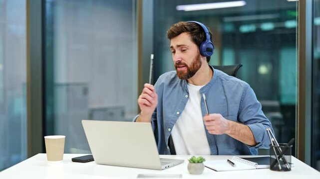 Excited businessman with headphones dances and sings during video conference on laptop in office. Joyful man celebrates success at workplace.