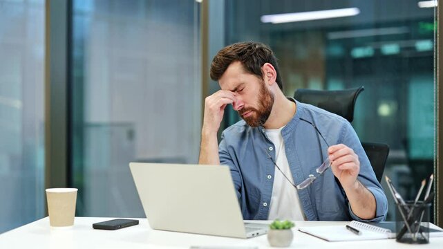 Tired businessman takes off glasses and rubs eyes from eyestrain while working on laptop in modern office. He experiences discomfort and fatigue.