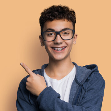 Excited happy black curly haired man in braces brackets, wear eye glasses, hoodie jacket advertise show pointing finger sales slogan text space, isolated brown background. Dental care ad. Square image
