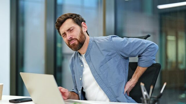 Young businessman with a beard in casual clothes sits in a chair at his desk in the office, working at a computer and experiences severe back pain.