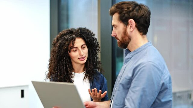 Two colleagues collaborate on a project, utilizing a laptop in a well-lit modern office. Professional woman presenting information to a bearded businessman.