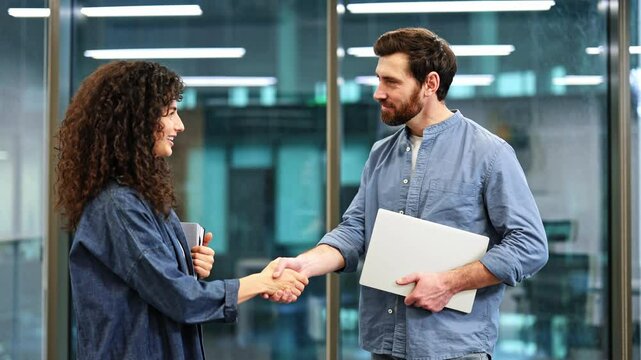 Two colleagues, one male and one female, make eye contact, smile, shake hands, and agree on something as they stand in an office.