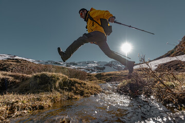 A man in bright clothes jumps with sticks against the background of snowy mountains and the sky. A tourist jumps over a stream in bright clothes, side view, the sun is visible.
