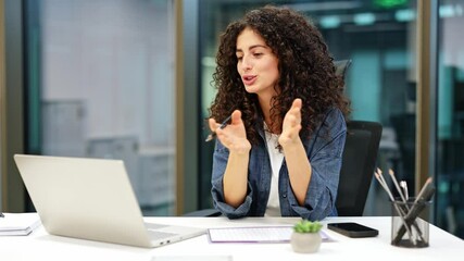 Confident curly-haired businesswoman working at modern glass office. Young female using laptop for video call. Business, technology, communication concept. - Powered by Adobe