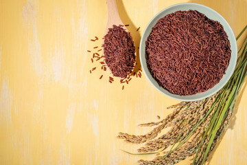 A bowl of red-rice is placed on a table next to a wood spoonful and ears of rice