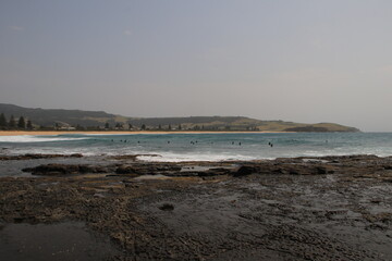 A serene coastal view showcasing a rocky shoreline with gentle ocean waves rolling in under a soft, hazy sky. Distant landforms add depth to the scene, creating a tranquil ambiance