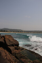 A serene coastal view showcasing a rocky shoreline with gentle ocean waves rolling in under a soft, hazy sky. Distant landforms add depth to the scene, creating a tranquil ambiance