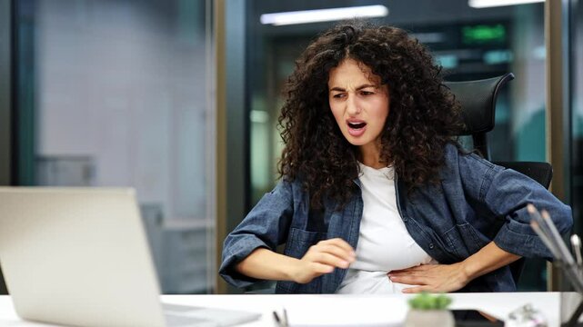 Curly haired businesswoman working in office at laptop having stomach or menstrual pain. She holds her hands to her belly and writhes in pain.
