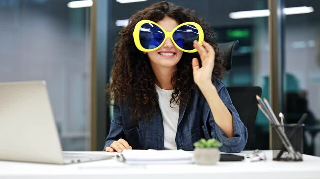 Happy businesswoman with curly hair wearing large funny glasses at modern office desk. Playful office worker enjoys a break, feels joy, and expresses positive emotions.