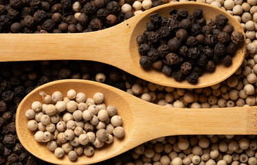 White and black peppercorns in wooden spoons, respectively, placed on peppercorns and photographed from above.