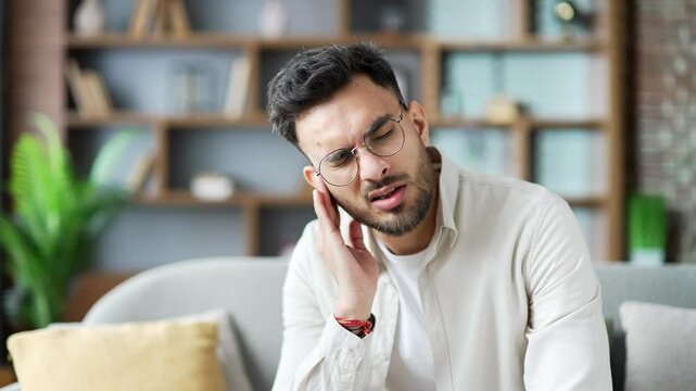 Earache. Sick man suffers from ear pain sitting on sofa in living room at home. Upset male touches his ear with his fingers. He has furuncle, shingles, eczema, ear diseases or otitis media. Close up