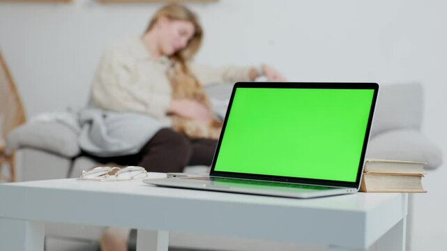 Laptop With Green Mock Up Screen On Table In Living Room. Background- Woman and Her Pet Dog Sitting on Sofa