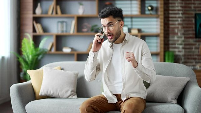 Angry excited man talking arguing on smartphone sitting on sofa in apartment at home. Frustrated young man shouting on phone call, expressing stress and anger emotions in modern living room interior