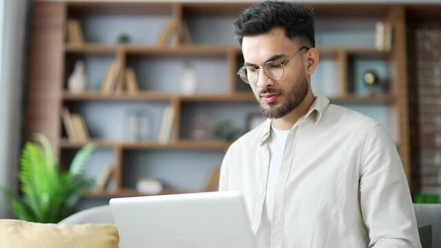 Handsome man working typing on a laptop sitting on ofa in living room at home office. Male freelancer using computer, texting a client, chatting online. busy with project looking at camera. Close up