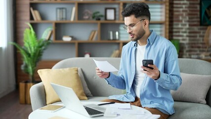 Happy man uses mobile app for spending bills using smartphone on phone while sitting at home in room. A male calculates household finances and enters data into a chat bot easy and simple to pay - Powered by Adobe