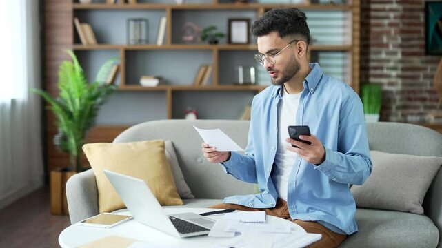 Confused young man having difficulty with paperwork while using mobile app for spending bills on phone sitting on sofa in living room at home office. Puzzled male cannot understand the problem